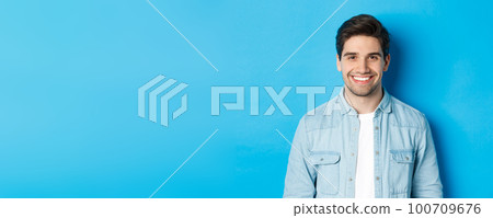 Close-up of young successful man smiling at camera, standing in casual outfit against blue background 100709676