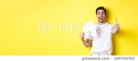 Real estate concept. Happy young male buyer showing thumb up and paper house model, smiling satisfied, standing over yellow background 100709703