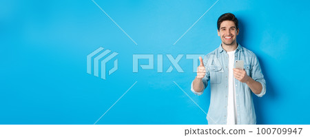 Concept of online shopping, applications and technology. Satisfied man in casual clothes smiling, showing thumbs up after using smartphone app, standing over blue background Concept of online shopping, applications and technology. Satisfied man in casual clothes smiling, showing thumbs up after using smartphone app, standing over blue background 100709947