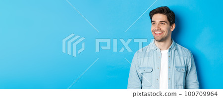 Young modern guy with beard, looking left and smiling pleased at copy space, standing over blue background 100709964