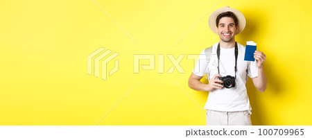 Travelling, vacation and tourism concept. Smiling man tourist holding camera, showing passport with tickets, standing over yellow background Travelling, vacation and tourism concept. Smiling man tourist holding camera, showing passport with tickets, standing over yellow background 100709965