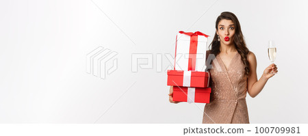 New Year, Christmas and celebration concept. Elegant woman in luxury dress and red lipstick, holding gifts and drinking champagne on party, standing over white background New Year, Christmas and celebration concept. Elegant woman in luxury dress and red lipstick, holding gifts and drinking champagne on party, standing over white background 100709981