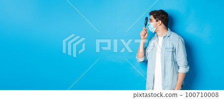 Concept of covid-19, social distancing and quarantine. Young man in medical mask searching for something, looking left through magnifying glass, standing against blue background Concept of covid-19, social distancing and quarantine. Young man in medical mask searching for something, looking left through magnifying glass, standing against blue background 100710008