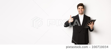 Portrait of confident businessman in black suit, holding clipboard with documents and showing thumb-up in approval, praise good job, standing against white background Portrait of confident businessman in black suit, holding clipboard with documents and showing thumb-up in approval, praise good job, standing against white background 100710034