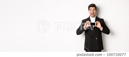 Image of cheerful, handsome man in black suit, showing smarthone screen and looking amazed, standing against white background Image of cheerful, handsome man in black suit, showing smarthone screen and looking amazed, standing against white background 100710077