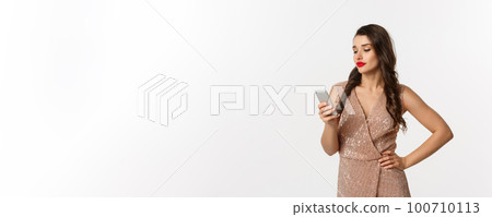 Christmas party and celebration concept. Careless woman in stylish dress reading message on mobile phone, looking skeptical, standing over white background 100710113