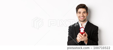 Image of handsome man looking romantic, open small box with engagement ring, making a proposal and smiling, standing against white background 100710322