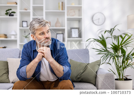 Adult mature man pensive sitting alone at home on sofa close up, gray haired person thinks looks towards window, depressed sad. 100710373