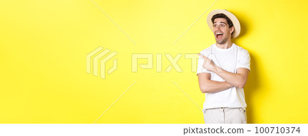 Concept of tourism and lifestyle. Excited handsome guy in straw hat checking out advertisement, pointing and looking at upper left corner logo, yellow background Concept of tourism and lifestyle. Excited handsome guy in straw hat checking out advertisement, pointing and looking at upper left corner logo, yellow background 100710374