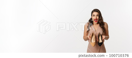 Party and celebration concept. Excited woman holding two types of heels and looking amazed, picking outfit for Christmas, standing in elegant dress over white background 100710376