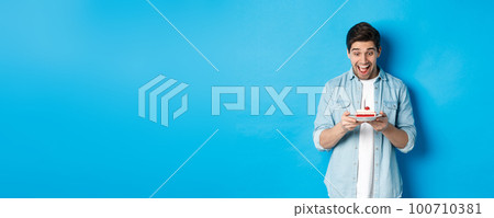 Excited young male model looking at birthday cake and smiling, making a wish and celebrating, standing against blue background 100710381