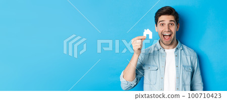 Real estate concept. Happy young man looking excited, found apartment, showing small house model, standing over blue background Real estate concept. Happy young man looking excited, found apartment, showing small house model, standing over blue background 100710423