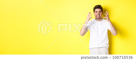 Angry man looking mad, grimacing and shaking hands furious, standing outraged against yellow background 100710536