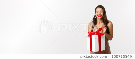 Merry Christmas. Charming woman in elegant dress open gift box and smiling delighted, unwrapping a present, standing over white background Merry Christmas. Charming woman in elegant dress open gift box and smiling delighted, unwrapping a present, standing over white background 100710549
