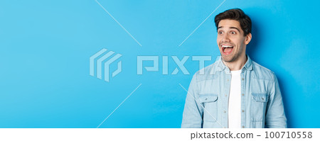 Close-up of handsome caucasian male model looking amazed left, checking out advertisement, standing over blue background Close-up of handsome caucasian male model looking amazed left, checking out advertisement, standing over blue background 100710558