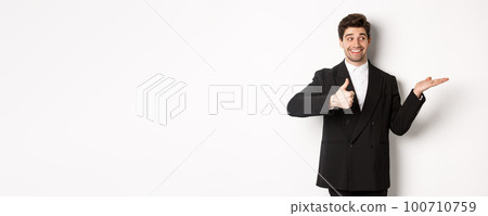 Handsome businessman in black suit, showing thumb-up and holding your product in hand over white copy space, standing against white background 100710759
