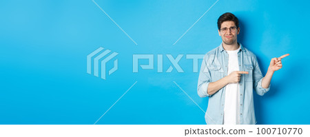 Disappointed young man in glasses pointing fingers right at copy space, showing promo banner and smirking dissatisfied, judging bad product, standing over blue background Disappointed young man in glasses pointing fingers right at copy space, showing promo banner and smirking dissatisfied, judging bad product, standing over blue background 100710770