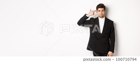 Portrait of distressed and troubled businessman in suit, pointing finger gun at head and looking bothered, shooting himself, standing against white background 100710794