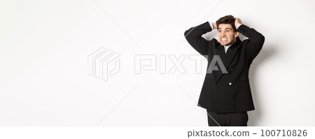 Image of frustrated and angry businessman in black suit, ripping hair on head and grimacing mad, looking left at disaster, standing tensed against white background 100710826