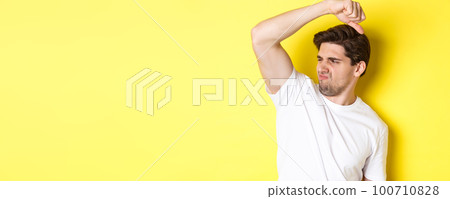 Man in sweat smelling his armpit, standing in white t-shirt and grimacing from stinky clothes 100710828