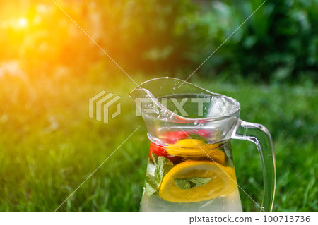 Defocus glass jug of lemonade with slice lemon and mint on natural green background with water drops. Pitcher of fresh cold ice summer cocktail or punch. Drops, wet. Copy space. Sunflare Out of focus Defocus glass jug of lemonade with slice lemon and mint on natural green background with water drops. Pitcher of fresh cold ice summer cocktail or punch. Drops, wet. Copy space. Sunflare Out of focus 100713736