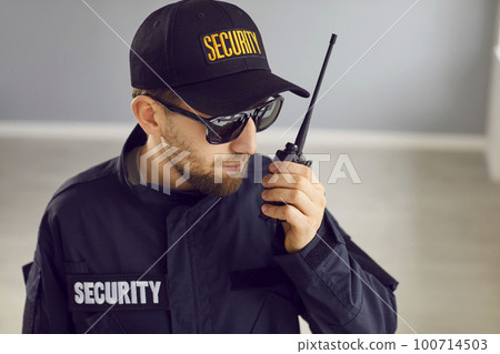 Young male security guard worker in black uniform monitoring house and using radio Young male security guard worker in black uniform monitoring house and using radio 100714503