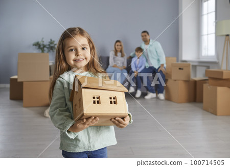 Happy child girl with toy wooden house in hands standing in new apartment with family in background. Happy child girl with toy wooden house in hands standing in new apartment with family in background. 100714505
