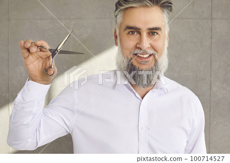 Portrait of a mature man barber with a grey beard and a mustache holding scissors and smiling Portrait of a mature man barber with a grey beard and a mustache holding scissors and smiling 100714527