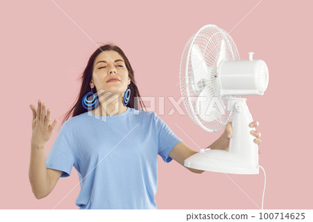 Happy young woman holding electric fan and breathing cool, fresh air blowing in her face 100714625
