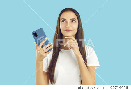 Happy beautiful young woman is holding her mobile phone, looking away and thinking 100714636