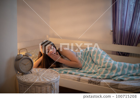 A tired woman yawns while lying on her bed covered with a soft blanket. 100715208