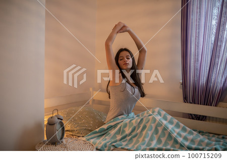 A woman stretches sitting on her bed in the bedroom. 100715209
