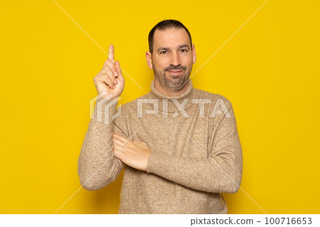 Bearded hispanic man wearing a beige turtleneck raising his index finger asking for a turn to express the great idea he had, he feels proud for having found a solution to the problem. Bearded hispanic man wearing a beige turtleneck raising his index finger asking for a turn to express the great idea he had, he feels proud for having found a solution to the problem. 100716653