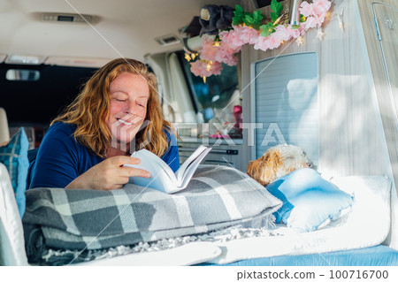 Middle-aged woman relaxing, reading book while lying in camper car vehicle with cockapoo puppy pet during road trip. Best friend. Enjoying free lifestyle, travel, vacation, freedom. Selective focus. 100716700
