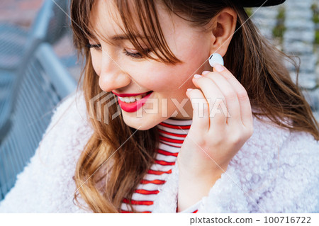Close up of a stylish young smiling woman using white true wireless earbuds by hand to put in ear and control. Putting on earbud into the ear for listening to music and podcasts. Selective focus. Close up of a stylish young smiling woman using white true wireless earbuds by hand to put in ear and control. Putting on earbud into the ear for listening to music and podcasts. Selective focus. 100716722