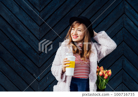 Portrait of young smiling fashion woman with reusable coffee cup and net bag with fresh tulip flowers squinting at the sun on the black wooden background. Urban city street fashion. Spring mood. 100716735