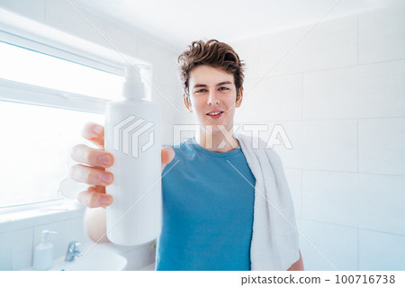 Smiling caucasian teenage boy with acne problem holding a white plastic mockup bottle for face skin care cosmetics at home. Teenager skin care everyday treatment process. Selective focus, copy space Smiling caucasian teenage boy with acne problem holding a white plastic mockup bottle for face skin care cosmetics at home. Teenager skin care everyday treatment process. Selective focus, copy space 100716738