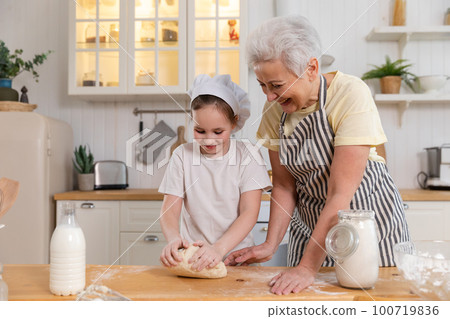 Happy family in kitchen. Grandmother and granddaughter child cook in kitchen together. Grandma teaching kid girl knead dough bake cookies. Household teamwork helping family generations concept 100719836