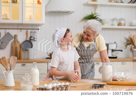 Happy family in kitchen. Grandmother and granddaughter child cook in kitchen together. Grandma teaching kid girl knead dough bake cookies. Household teamwork helping family generations concept 100719876