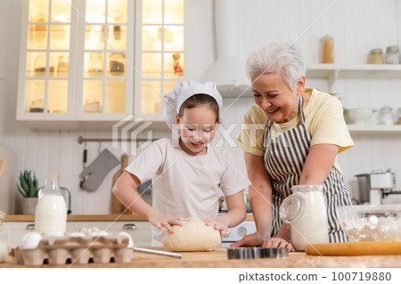 Happy family in kitchen. Grandmother and granddaughter child cook in kitchen together. Grandma teaching kid girl knead dough bake cookies. Household teamwork helping family generations concept Happy family in kitchen. Grandmother and granddaughter child cook in kitchen together. Grandma teaching kid girl knead dough bake cookies. Household teamwork helping family generations concept 100719880