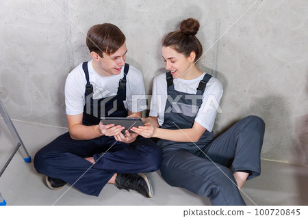 House DIY renovation concept. Young happy couple sitting on floor looking home repair plan in tablet indoors. Woman showing to man plan interior design online. Couple in new home during repair works House DIY renovation concept. Young happy couple sitting on floor looking home repair plan in tablet indoors. Woman showing to man plan interior design online. Couple in new home during repair works 100720845