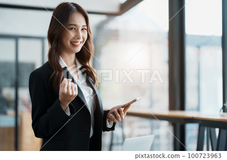 Business work concept, portrait of a young businesswoman holding a cell phone showing a happy expression, business success Business work concept, portrait of a young businesswoman holding a cell phone showing a happy expression, business success 100723963