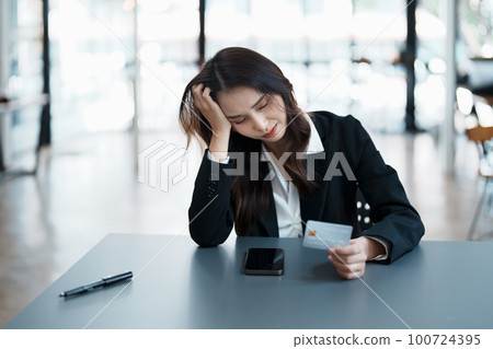 Portrait of young Asian woman holding credit card with smart phone mobile and showing worried expression on credit card debt 100724395