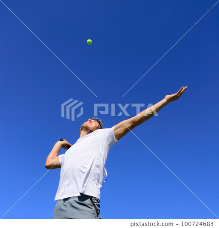 Tennis player man doing serve in position to hit ball in the air. Playing tennis outdoors on blue sky summer background. Active sport athlete doing fitness fun. 100724683
