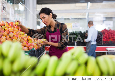 Woman in apron stacking plums in greengrocer 100725483