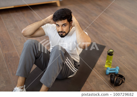 Indian man in sport clothes doing sit-ups in living room 100729682
