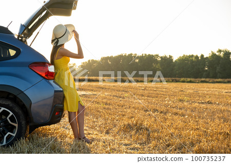 Young woman driver in yellow summer dress and straw hat standing near a blue car enjoying warm summer day at sunset. Young woman driver in yellow summer dress and straw hat standing near a blue car enjoying warm summer day at sunset. 100735237