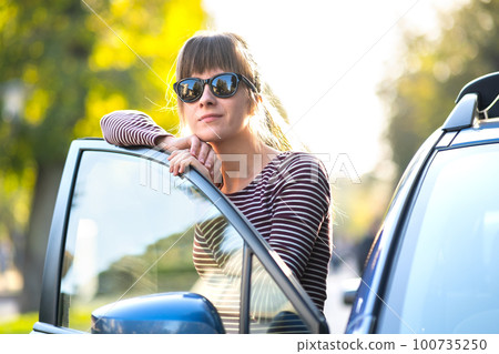 Young woman driver enjoying warm summer day standing beside her car on city street. Travelling and vacation concept. Young woman driver enjoying warm summer day standing beside her car on city street. Travelling and vacation concept. 100735250