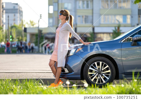 Young woman driver enjoying warm summer day standing beside her car on city street. Travelling and vacation concept. Young woman driver enjoying warm summer day standing beside her car on city street. Travelling and vacation concept. 100735251