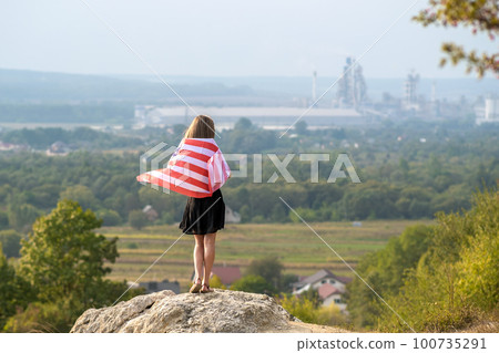 Young pretty american woman with long hair holding waving on wind USA flag on her sholders standing outdoors enjoying warm summer day. 100735291